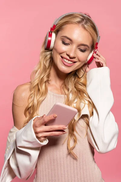 Belle femme blonde heureuse avec écouteurs en utilisant smartphone isolé sur rose — Photo de stock