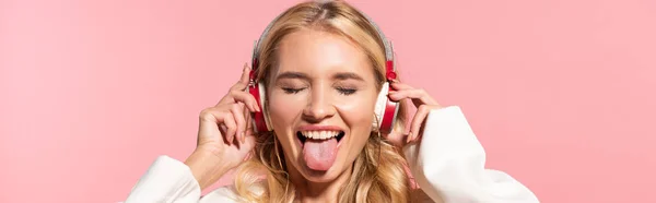 Panoramaaufnahme der schönen blonden Frau mit Kopfhörern und geschlossenen Augen, die die Zunge isoliert auf rosa — Stockfoto