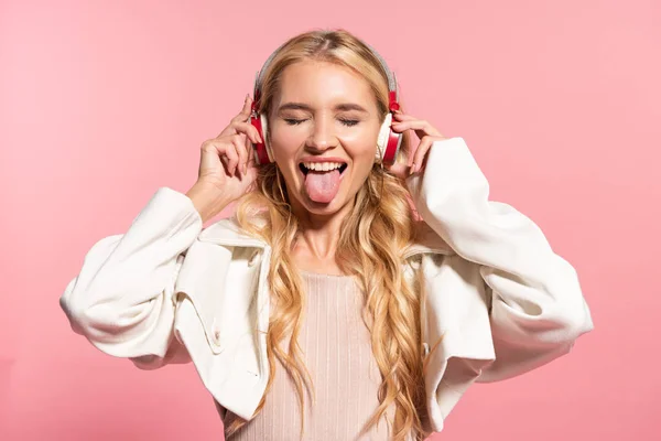 Красива блондинка з навушниками і закритими очима стирчить язиком ізольовано на рожевому — стокове фото