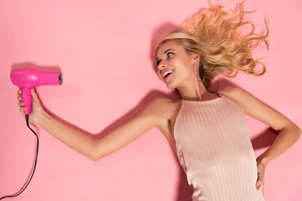 Smiling beautiful blonde woman lying and holding hairdryer on pink — Stock Photo