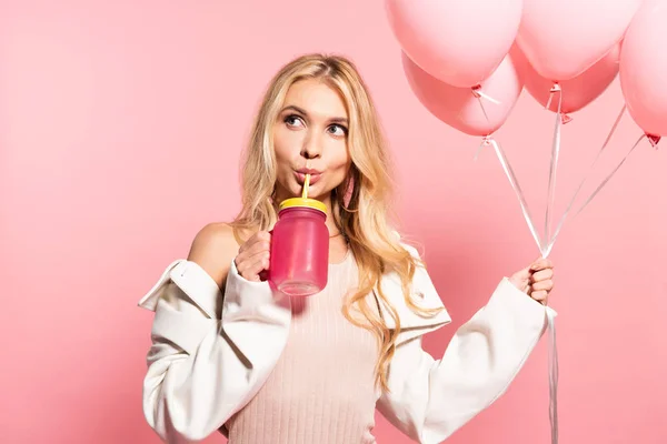 Hermosa mujer rubia bebiendo de paja y sosteniendo globos rosados en rosa - foto de stock