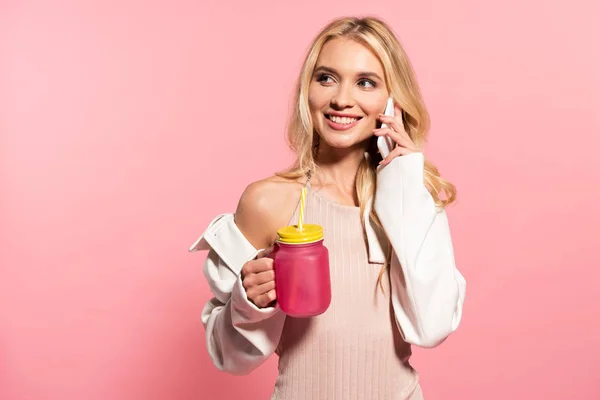 Hermosa chica rubia sosteniendo la bebida y hablando en el teléfono inteligente en rosa - foto de stock