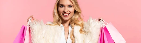 Feliz hermosa mujer rubia en chaqueta de piel sintética blanca sosteniendo bolsas de compras aisladas en rosa, tiro panorámico - foto de stock