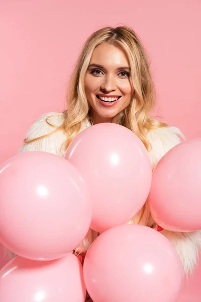 Happy beautiful blonde woman in faux fur jacket holding pink balloons isolated on pink — Stock Photo