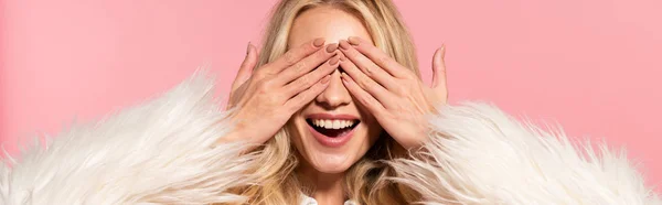Glücklich schöne blonde Frau in weißer Kunstpelzjacke mit Händen auf Augen isoliert auf rosa, Panoramaaufnahme — Stockfoto