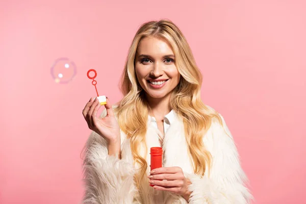 Happy blonde girl in white faux fur coat holding soap bubbles isolated on pink — Stock Photo