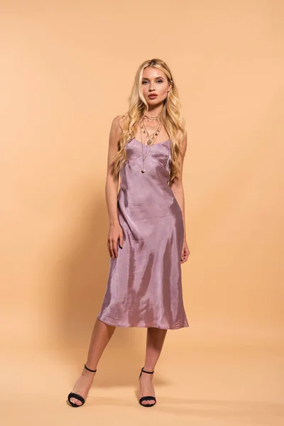 Elegante mujer rubia en vestido de satén violeta y collar posando en beige - foto de stock