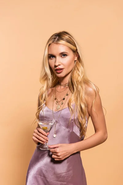 Elegant woman in violet satin dress and necklace holding cocktail isolated on beige — Stock Photo