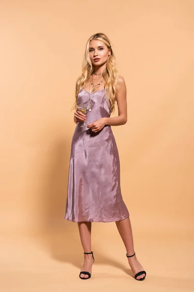 Elegant blonde woman in violet satin dress and necklace holding cocktail on beige — Stock Photo