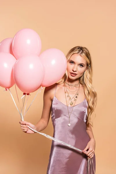 Elegante mujer rubia en vestido de satén violeta y collar sosteniendo globos rosados en beige - foto de stock