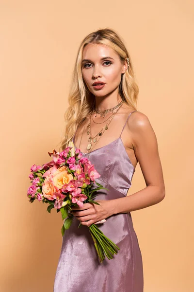 Elegante blonde Frau in violettem Satinkleid und Halskette mit Blumenstrauß auf Beige — Stockfoto