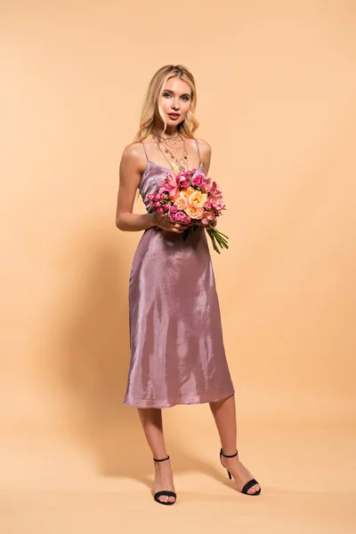 Elegante blonde Frau in violettem Satinkleid mit Blumenstrauß auf beige — Stockfoto