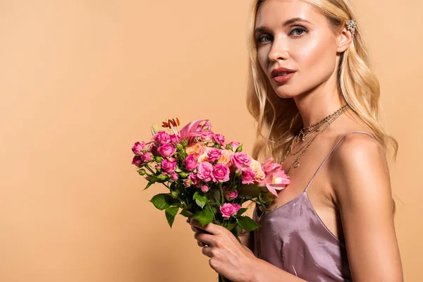 Blonde woman in violet satin dress and necklace holding bouquet of flowers on beige — Stock Photo