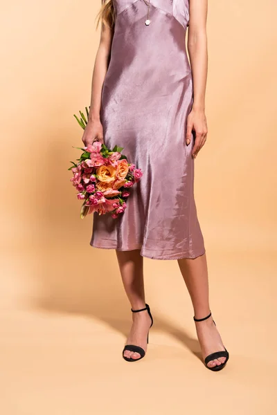 Vista ritagliata di donna elegante in abito di raso viola con mazzo di fiori su beige — Foto stock
