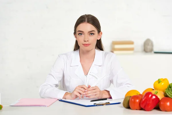 Front view of dietitian in white coat at workplace with vegetables, folder and clipboard on table — Stock Photo
