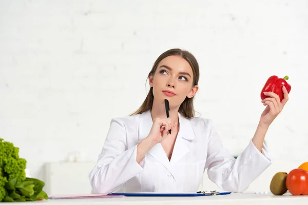 Nachdenkliche Diätassistentin im weißen Mantel mit rotem Paprika und Stift und Blick zum Arbeitsplatz — Stockfoto