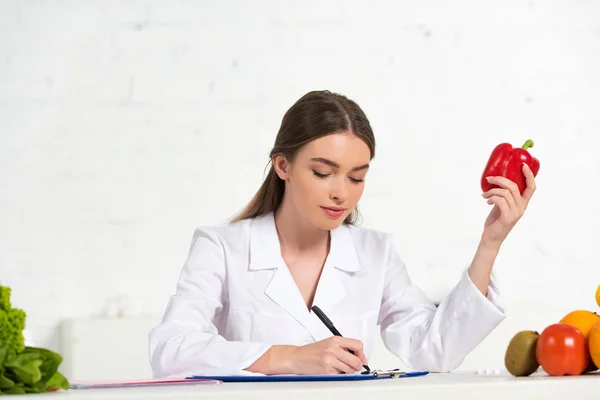 Dietista en bata blanca sosteniendo pimiento rojo y escribiendo en portapapeles en el lugar de trabajo - foto de stock