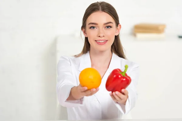 Vue de face d'une diététiste souriante en manteau blanc tenant du poivron orange et rouge — Photo de stock