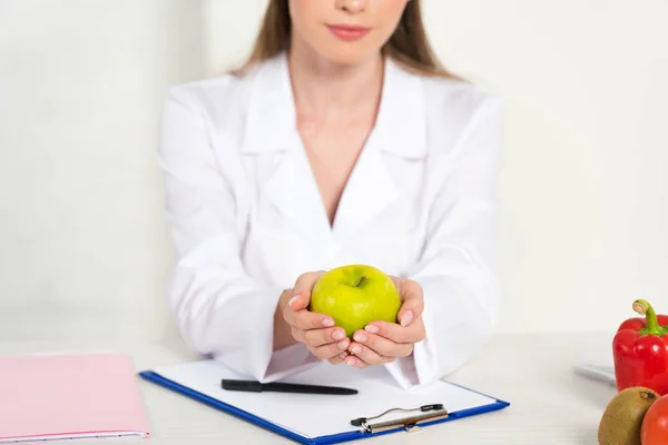 Vista parcial del dietista en capa blanca sosteniendo manzana en el lugar de trabajo - foto de stock