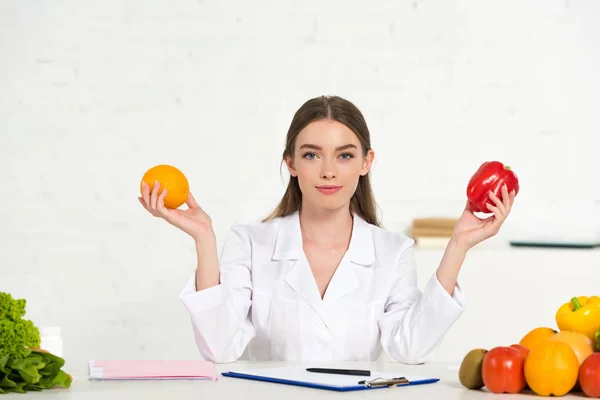 Vista frontal del dietista en bata blanca sosteniendo naranja y pimiento en el lugar de trabajo - foto de stock