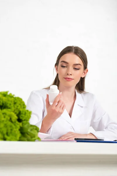 Pensive dietitian in white coat holding pills at workplace — Stock Photo