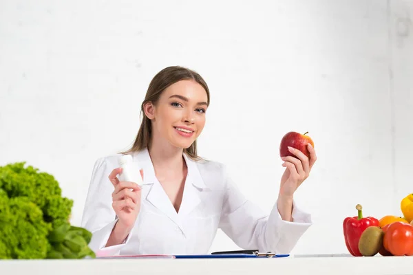 Smiling dietitian in white coat holding pills and apple at workplace — Stock Photo