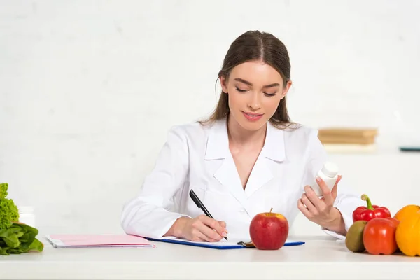 Lächelnde Diätassistentin in weißem Kittel mit Tabletten und Schriftzug am Arbeitsplatz mit Obst und Gemüse auf dem Tisch — Stockfoto