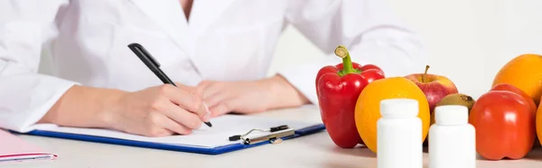 Panoramic shot of dietitian in white coat writing in clipboard at workplace with pills, fruits and vegetables on table — Stock Photo