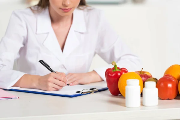 Cropped view dietitian in white coat writing in clipboard at workplace with pills, fruits and vegetables on table — Stock Photo