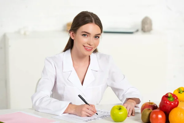 Улыбающийся диетолог в белом халате пишет на рабочем месте с фруктами и овощами на столе — стоковое фото