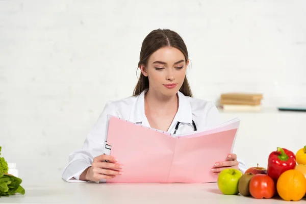 Konzentrierte Ernährungsberaterin in weißem Kittel liest Folder am Arbeitsplatz mit Obst und Gemüse auf dem Tisch — Stockfoto