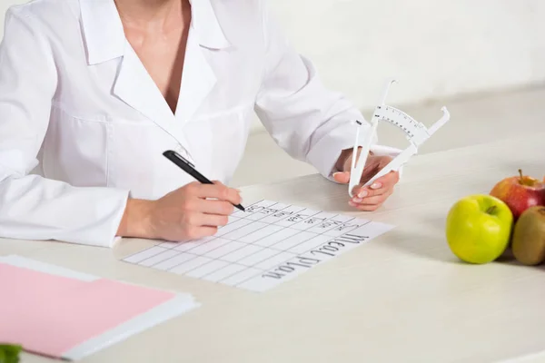 Partial view of dietitian holding caliper and writing meal plan at workplace with fruits on table — Stock Photo