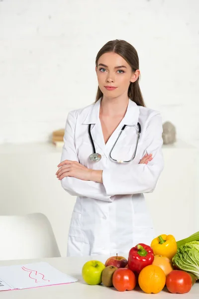 Atractivo dietista en bata blanca con equipo de pie con brazos cruzados cerca de la mesa con frutas y verduras frescas - foto de stock