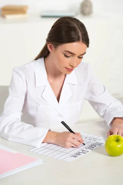 Focused dietitian in white coat writing meal plan at table with green apple — Stock Photo