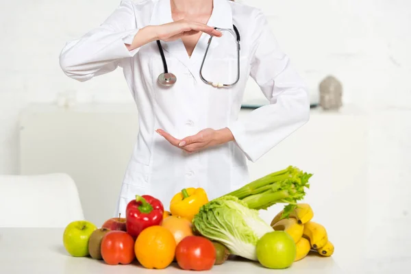 Teilansicht von Diätassistentin im weißen Mantel mit Stethoskop in der Nähe von frischem Obst und Gemüse — Stockfoto