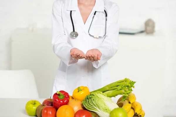 Vista parcial del dietista en bata blanca con estetoscopio cerca de frutas y verduras frescas - foto de stock