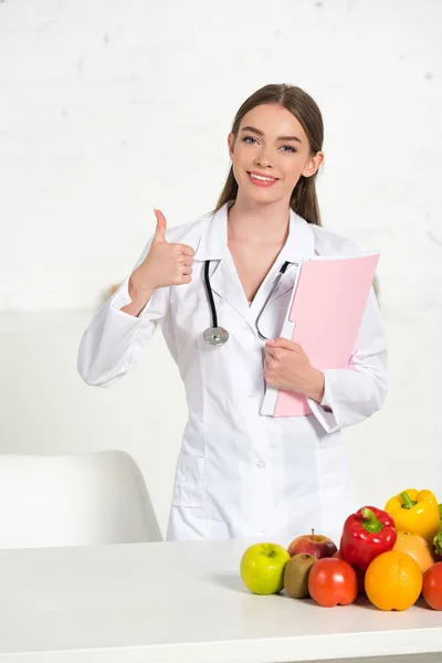 Dietista sonriente en la carpeta de la celebración del abrigo blanco y mostrando el pulgar hacia arriba cerca de la mesa con frutas y verduras frescas - foto de stock