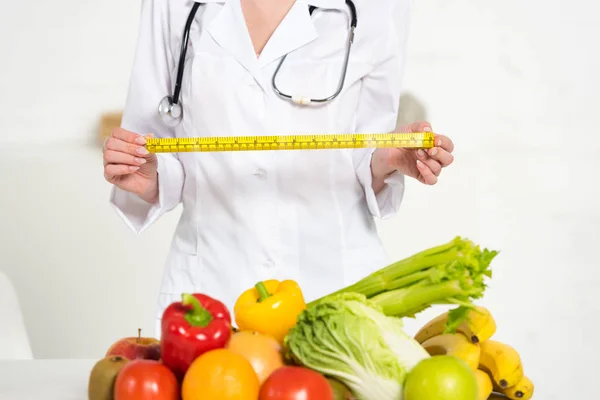 Cropped view of dietitian in white coat holding measure tape near fresh fruits and vegetables — Stock Photo