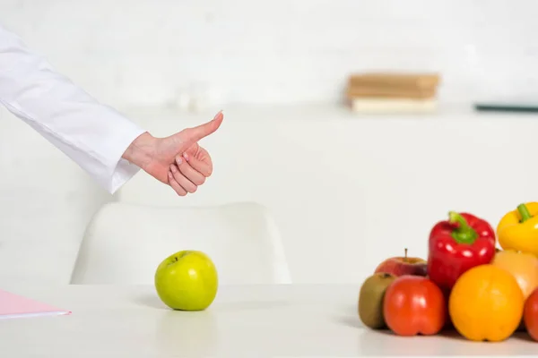 Cropped view of dietitian showing thumb up near table with fresh vegetables and fruits — Stock Photo