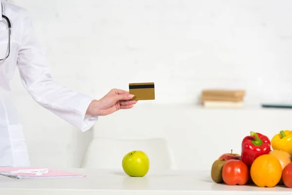 Vista recortada de dietista en bata blanca con tarjeta de crédito cerca de la mesa con frutas y verduras frescas - foto de stock
