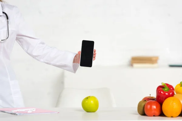 Vista ritagliata di dietista in bianco appendiabiti smartphone con schermo bianco vicino al tavolo con frutta e verdura — Foto stock