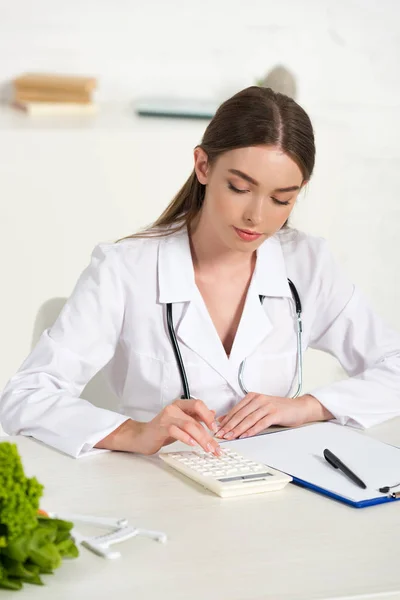 Focused dietitian in white coat with stethoscope using calculator at workplace — Stock Photo