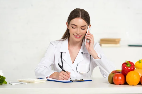 Dietista sonriente en bata blanca hablando en smartphone y escribiendo en portapapeles en el lugar de trabajo - foto de stock