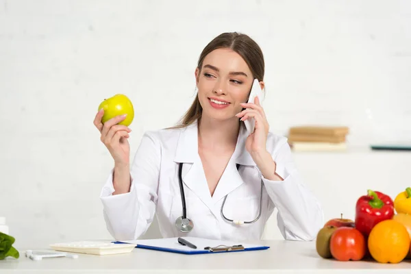 Dietista sonriente en bata blanca hablando en el teléfono inteligente y sosteniendo manzana en el lugar de trabajo - foto de stock