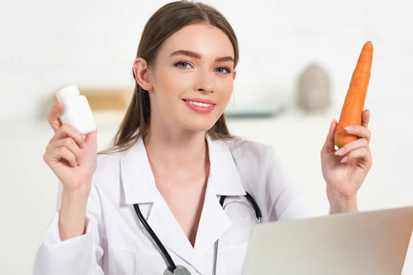 Smiling dietitian in white coat holding pills and carrot near laptop — Stock Photo