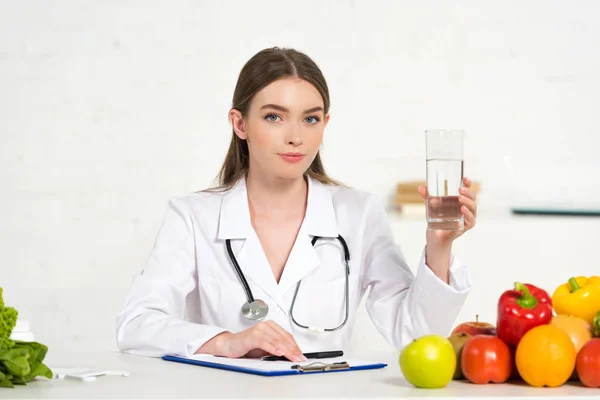 Dietitian in white coat holding glass of water at workplace — Stock Photo