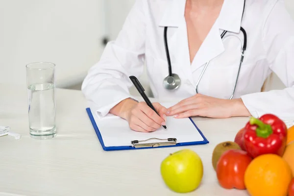 Vista recortada de nutricionista em jaleco branco escrevendo em prancheta no local de trabalho — Fotografia de Stock