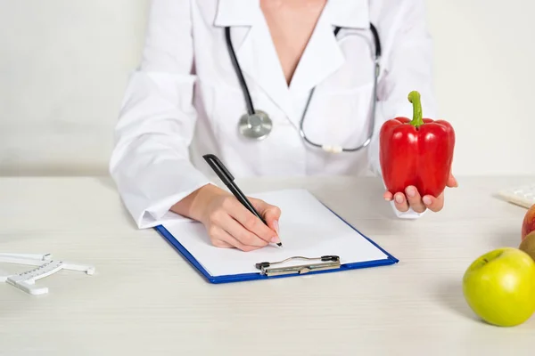 Cropped view of dietitian in white coat writing in clipboard and holding bell pepper at workplace — Stock Photo