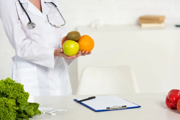 Partial view of dietitian in white coat holding fresh fruits near workplace — Stock Photo