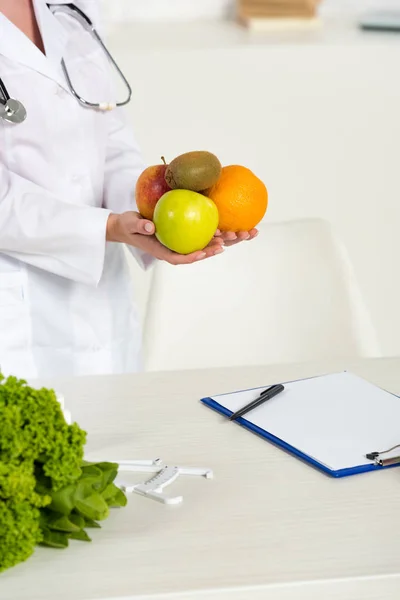 Vista parcial del dietista en bata blanca sosteniendo frutas frescas cerca del lugar de trabajo - foto de stock